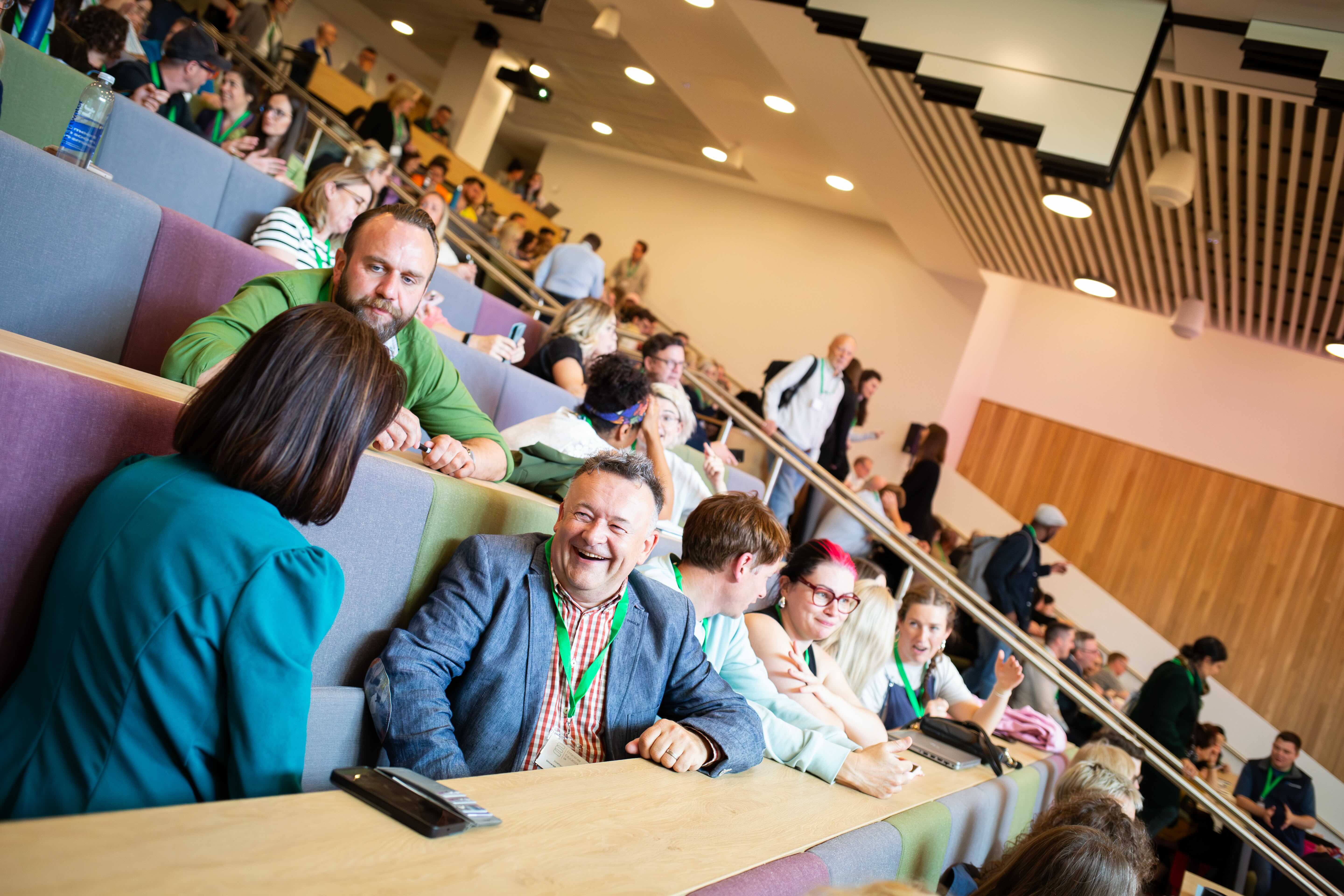 A lecture theatre with tiered seating full of happy and smiley conference delegates at IEEC2024.